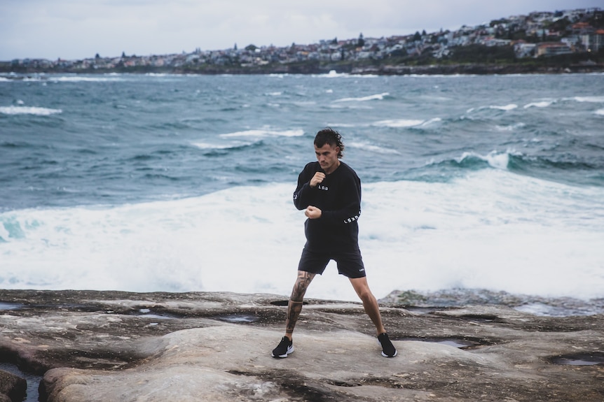 A boxer stands on rocks with fists prepped as if fighting, while behind him is the ocean with white caps visible in the water.