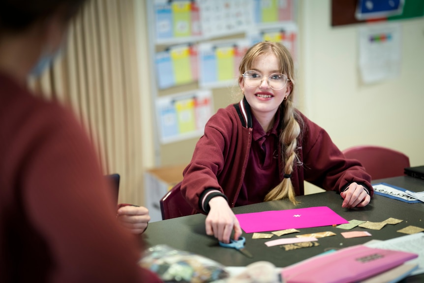 A teenage girl with blond hair in a long side plait sits, smiling, at a table doing an activity.