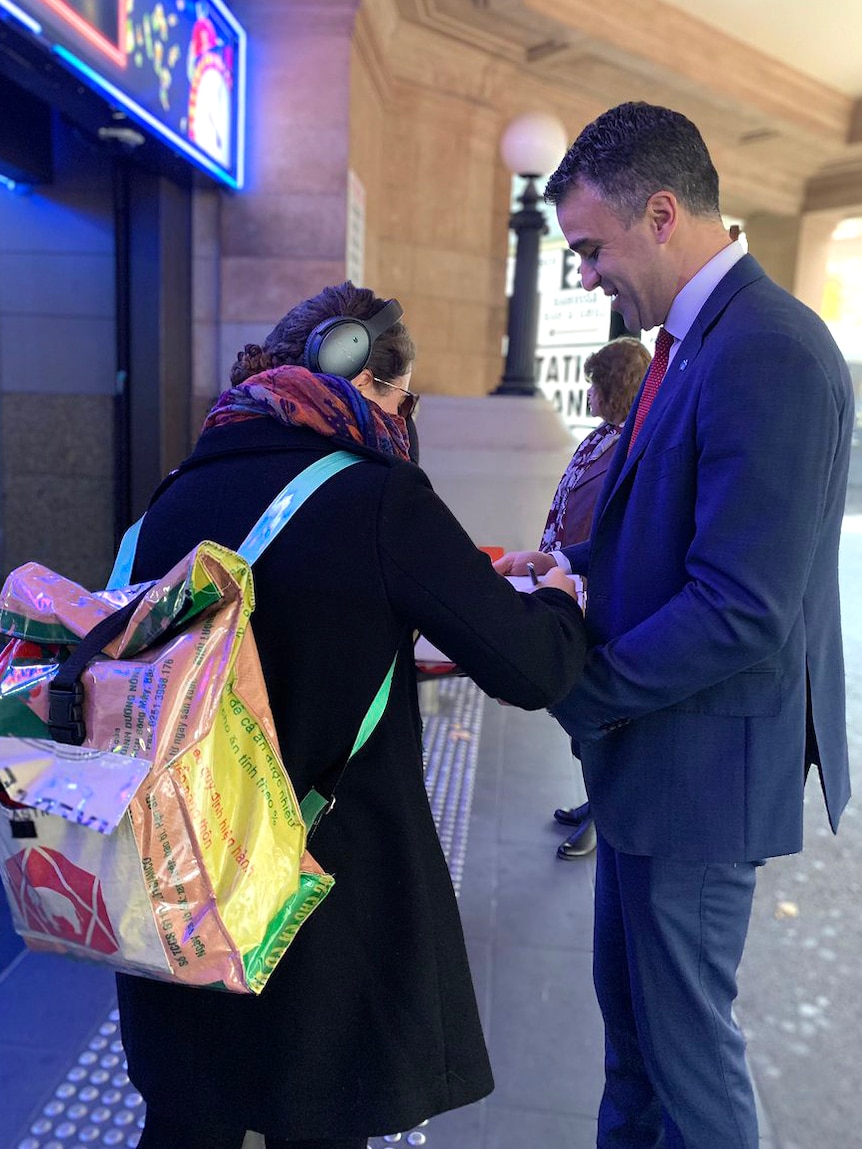 A man in a suit holds a clipboard being signed by a commuter at a railway station.