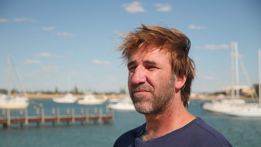 A man in a blue t-shirt looks out to sea, with boats and a pier in the background.