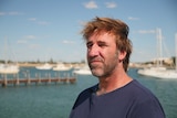 A man in a blue t-shirt looks out to sea, with boats and a pier in the background.