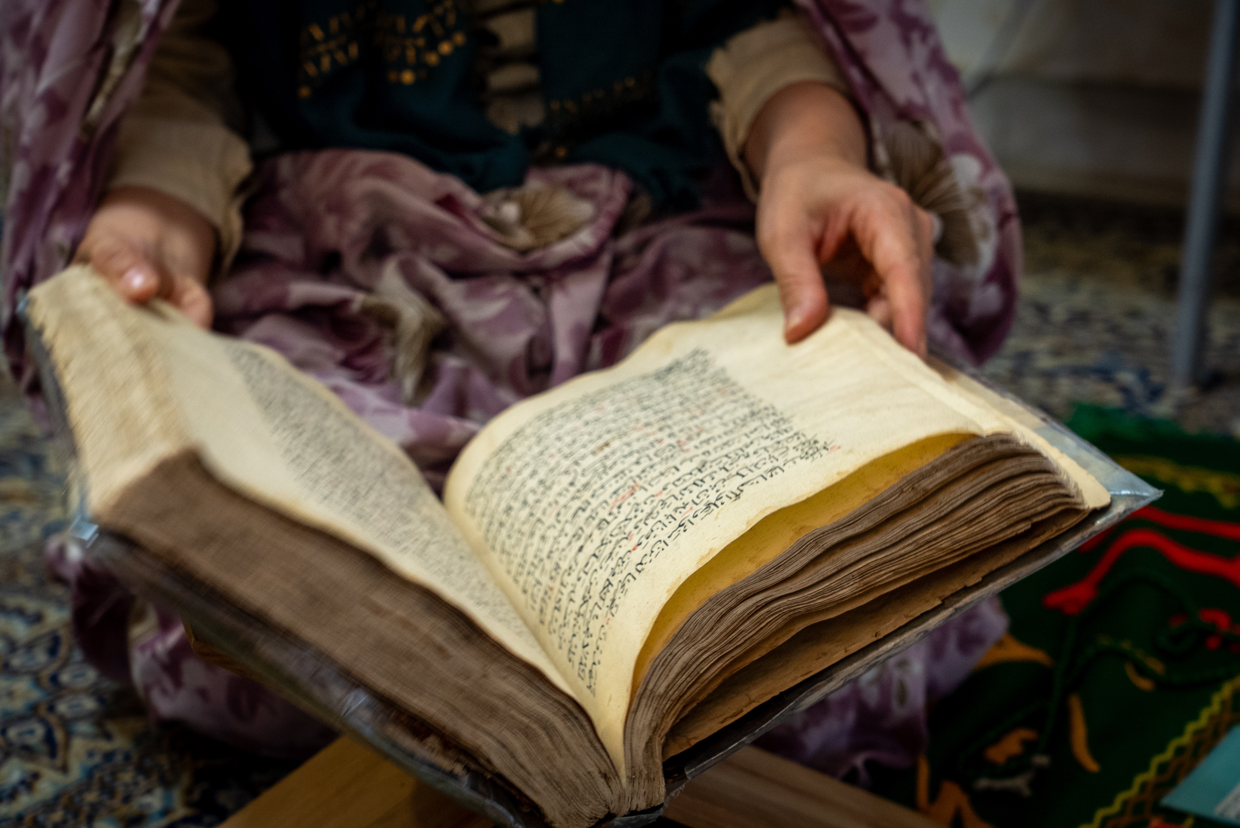 A woman reads a large book while sitting on the floor of a home.