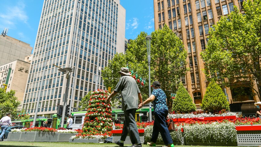 A couple walk hand in hand in Melbourne CBD with tall buildings, Christmas decorations and a tram passing by.