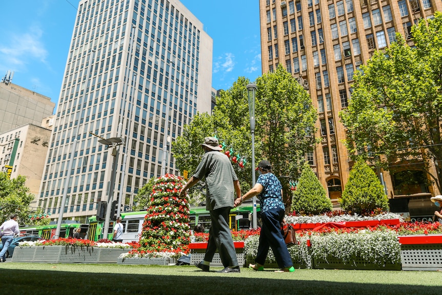 A couple walk hand in hand in Melbourne CBD with tall buildings, Christmas decorations and a tram passing by.
