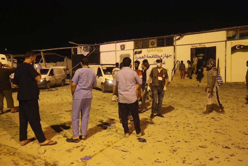 People stand outside a building. Small pieces of rubble appear to be on the ground.