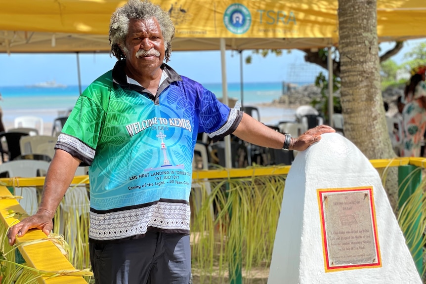 A Torres Strait Islander man with grey hair and blue and green shirt stands next to a stone memorial.