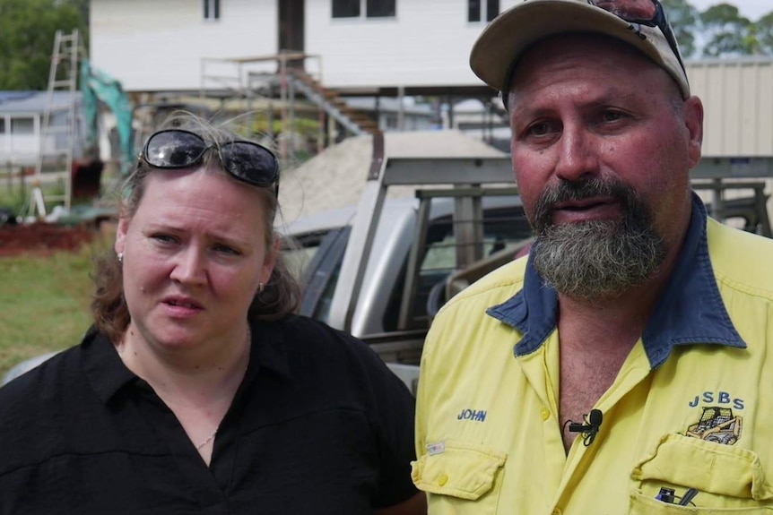A middle-aged woman in black stands alongside a man wearing a cap and worker's clothes at work site.