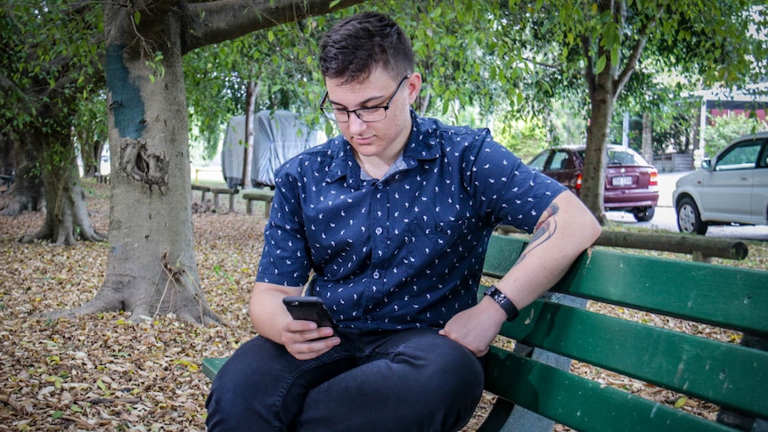 Transmasculine person Ash Polzin sits in a park reading a mobile phone.