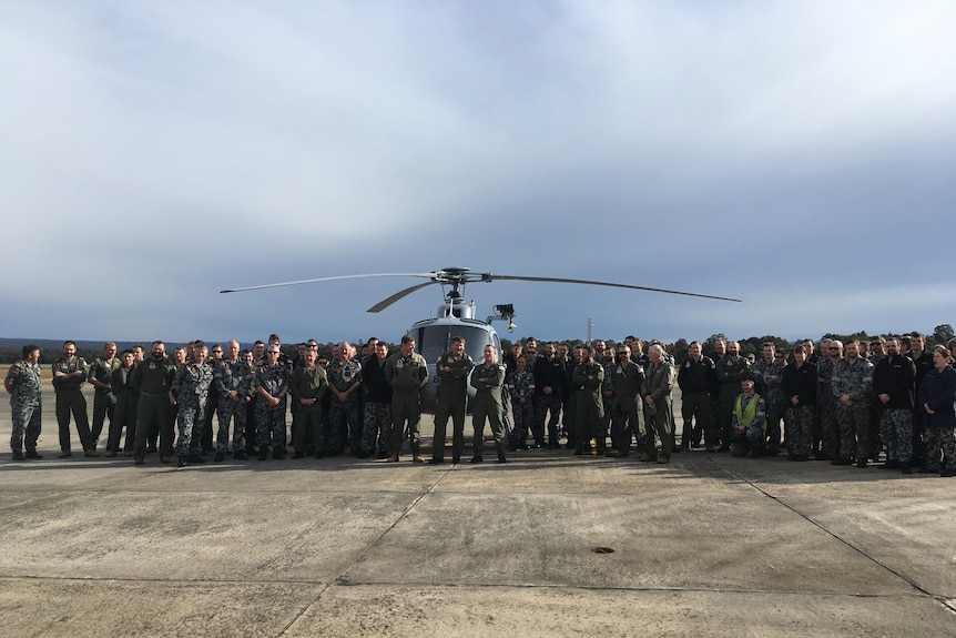 Navy squadron members stand on a tarmac in front of a grey helicopter.