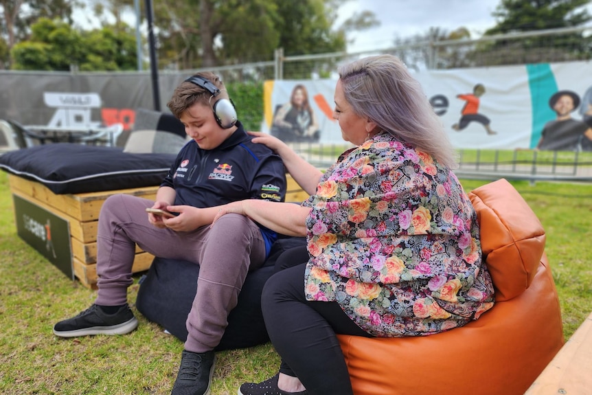 A boy with headphones looking at his phone while on a bean bag with his mum 