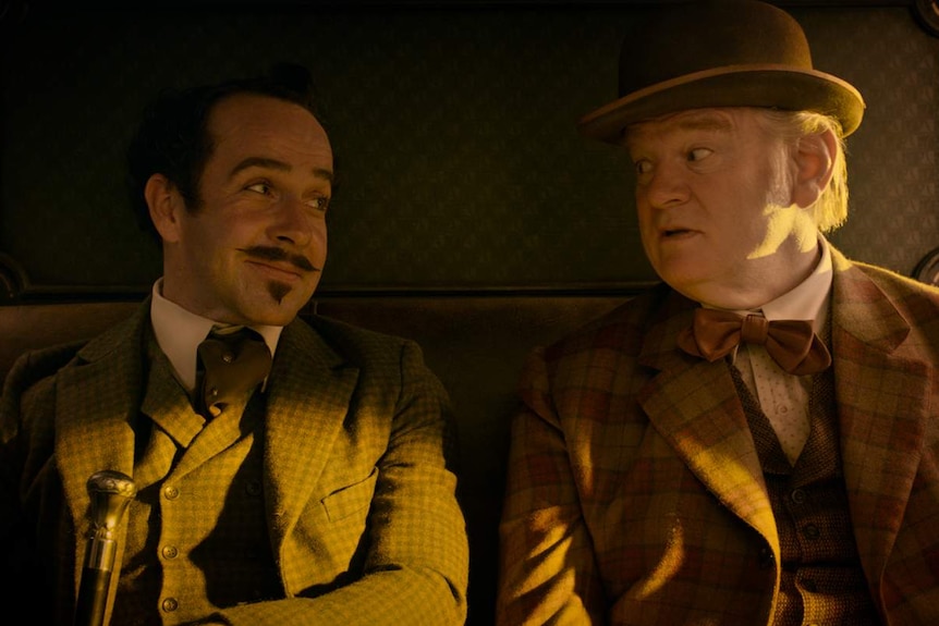 Two men dressed in suits sit inside a stage coach, looking at each other, one smiling.