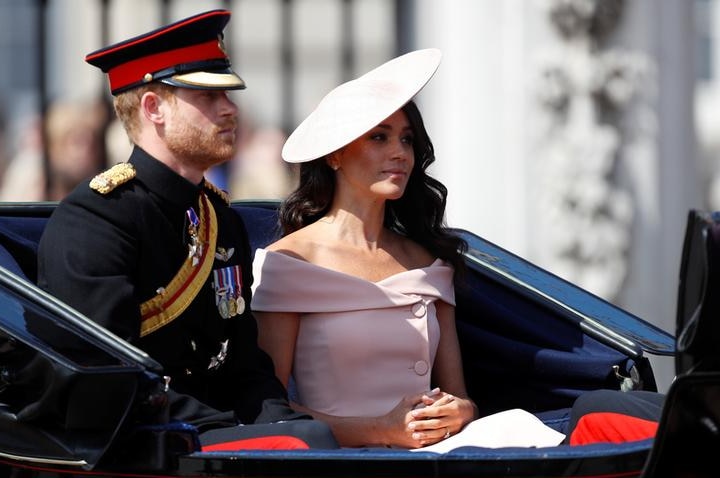 Prince Harry wears black military dress and cap as he sits in carriage next to Meghan, who wears a pale pink dress and hat.