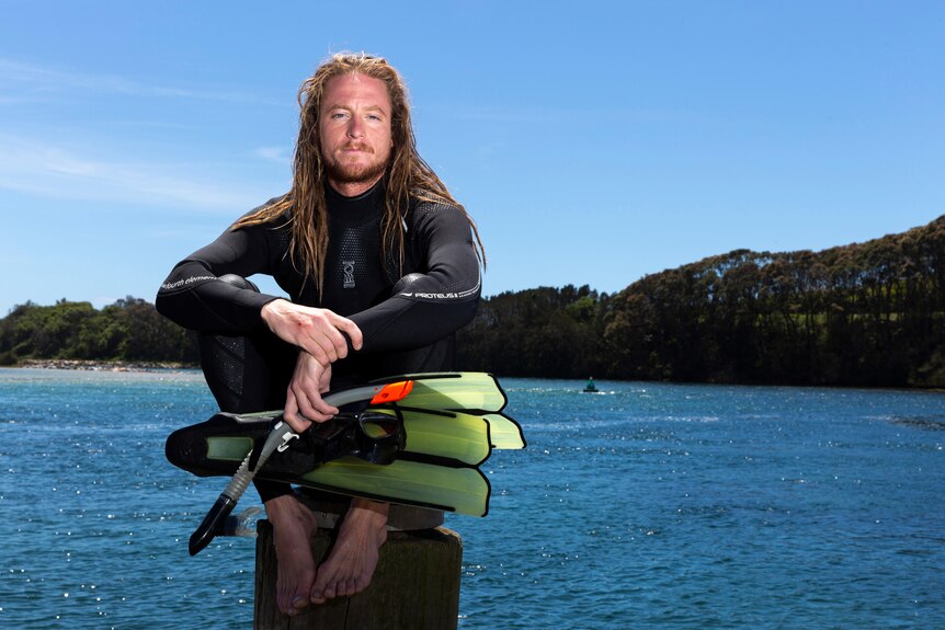 A young man in diving gear, perched up on a pylon by the ocean while looking at the camera