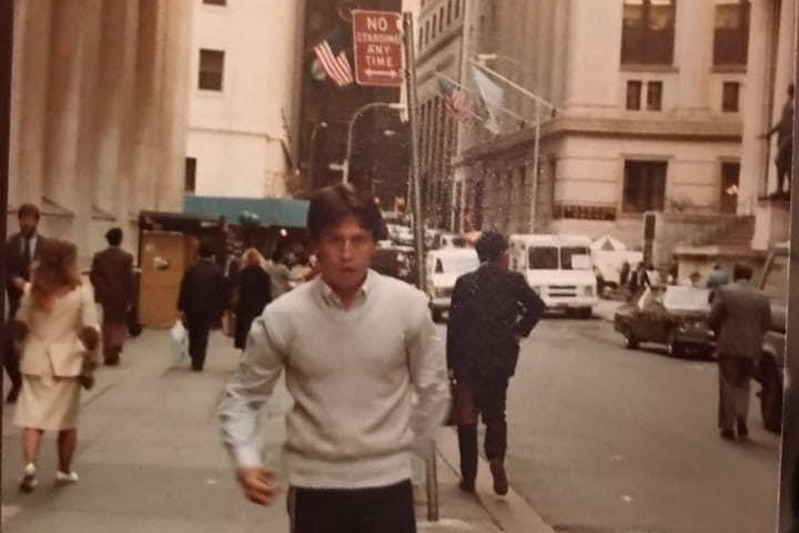 A young man with dark hair wearing a light sweater and dark pants stands in a New York street in the 1980s.