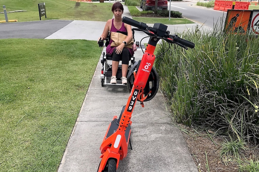 A woman in a wheelchair, with an e-scooter blocking her way on a footpath.