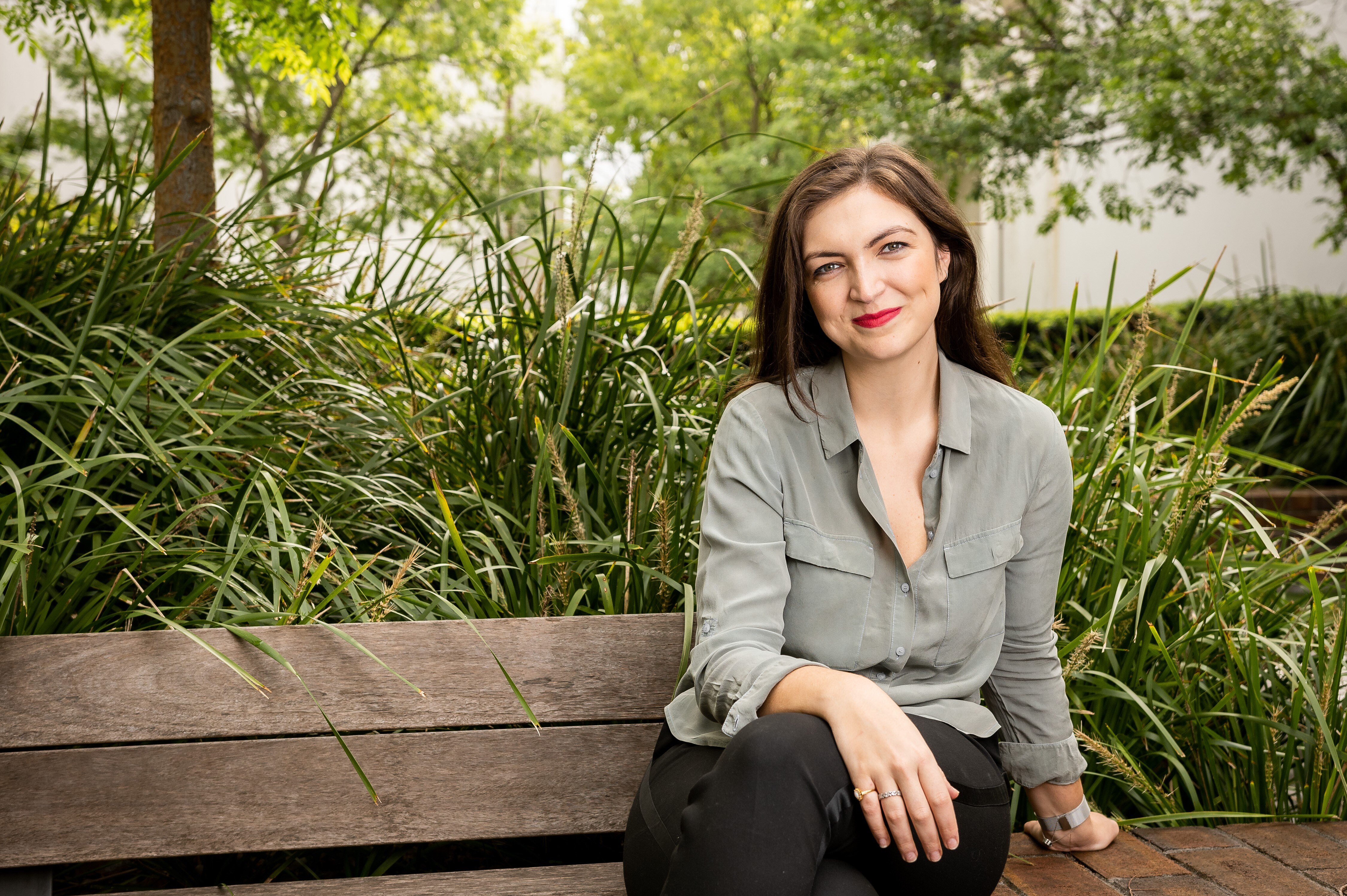 Aleks is wearing a green olive top, smiling and sitting on a park bench