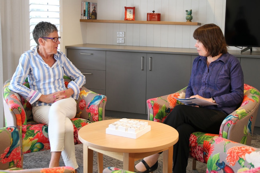 A woman with short grey hair and a woman with medium-length grey hair sitting in armchairs talking.