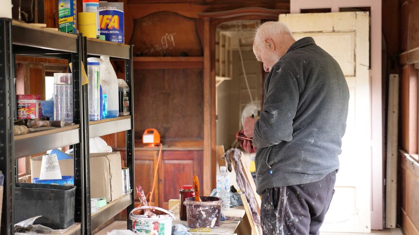 An old man with white facial hair in a grey jumper labels a tin can.