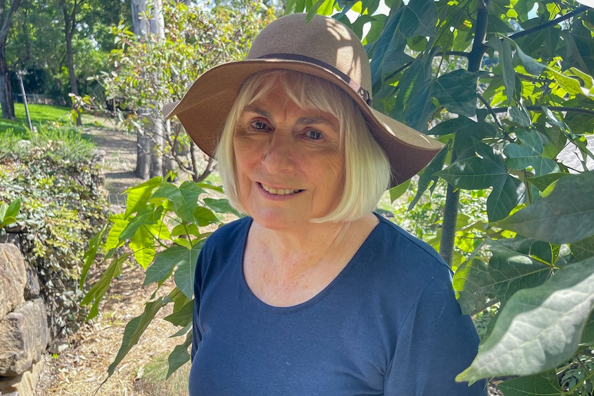 A woman in a blue shirt and tan felt hat standing in front of an Illawarra flame tree's green foliage.