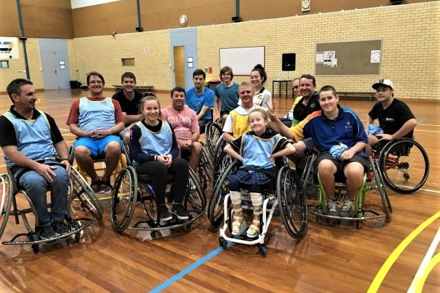 14 adults and kids in wheelchairs on basketball court, girls in leg braces in foreground