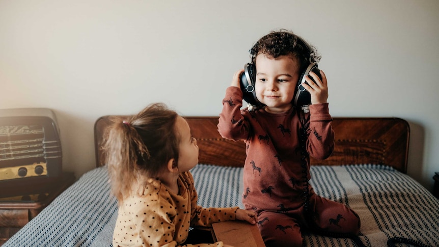 Children playing with headphones and a book on a bed, beside which sits a valve radio