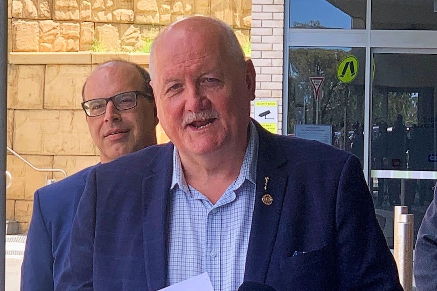 A man with a bald head and a moustache, wearing a suit, stands outside a building.