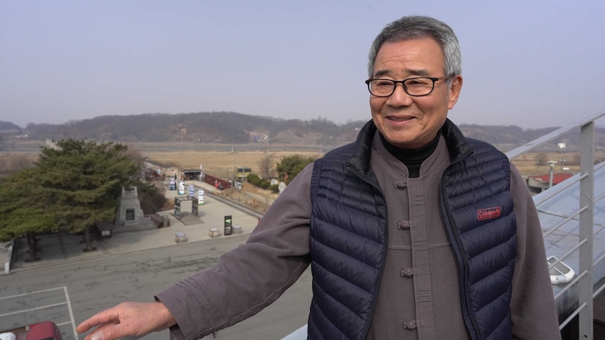 A man smiles at the camera standing in front of a military zone.