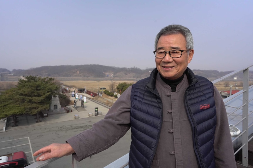 A man smiles at the camera standing in front of a military zone.