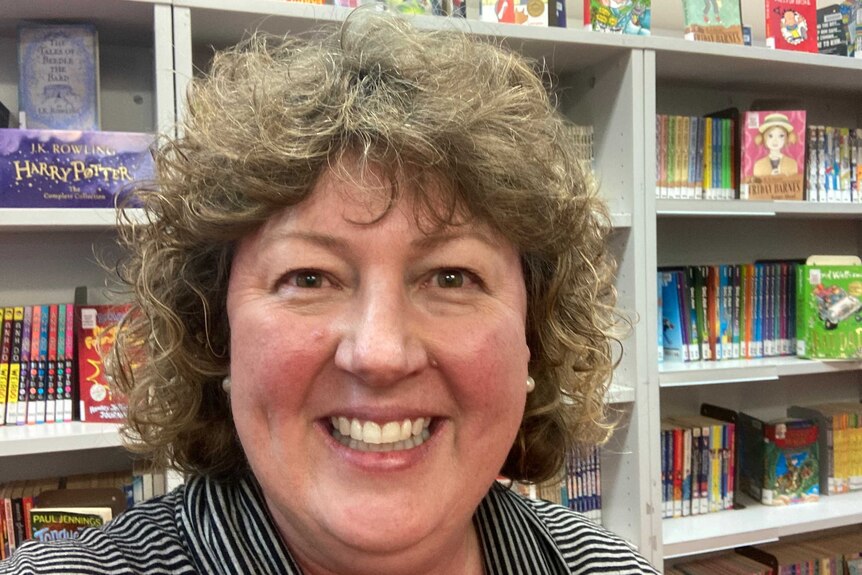 Davida McDonald has curly short hair and a striped shirt, she's in a library with shelves full of colourful children's books.