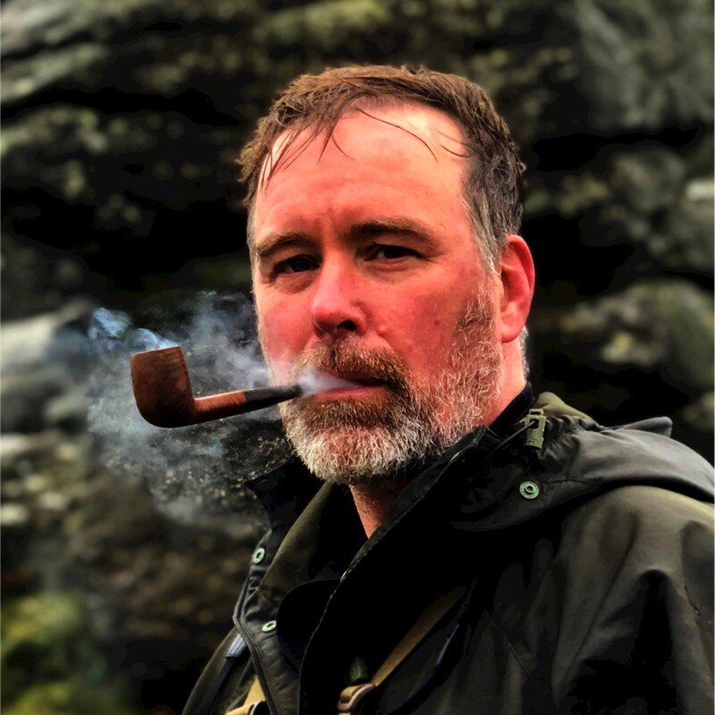 A flushed, bearded man smoking a pipe, apparently outdoors.