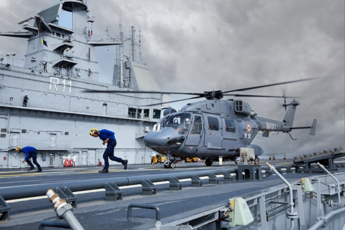 Two deck crew bend forward to keep heads low as they run from where a grey helicopter lands.