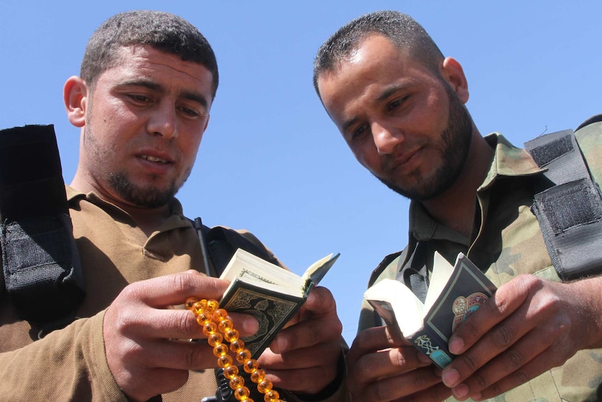 Two men in military uniforms look down at small Qurans they hold in their hands.