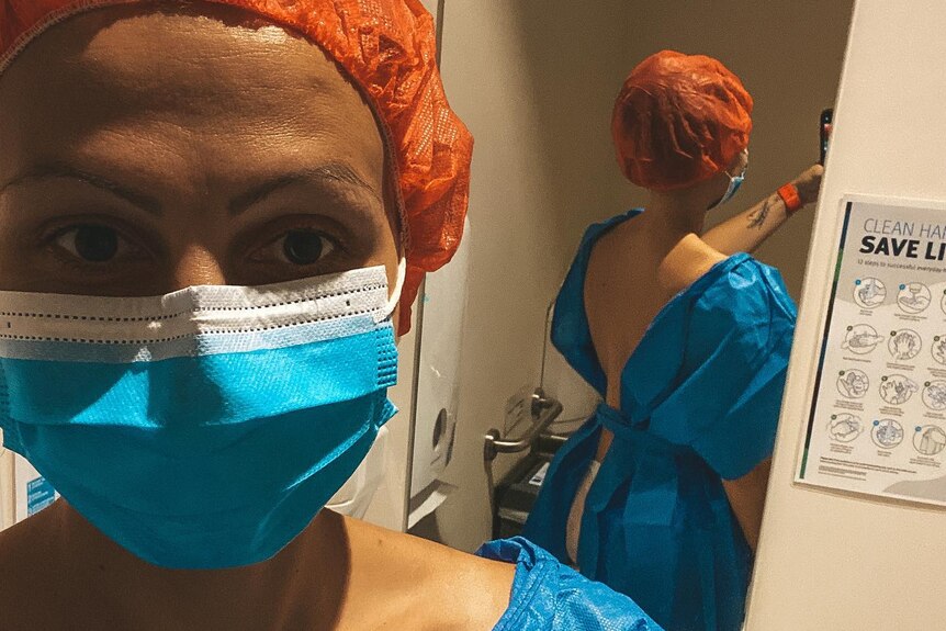 Maria in a blue surgical gown, blue surgical mask and orange surgical hair mask, taking a photo in a mirror.