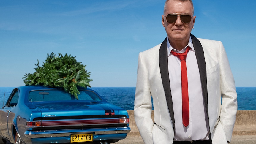 Jimmy Barnes sands in a carpark overlooking the beach he wears a suit and sunnies, car in the background has a christmas tree in
