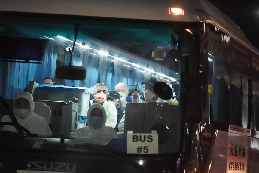 People on a bus wear surgical masks as the drivers wear hazard suits.