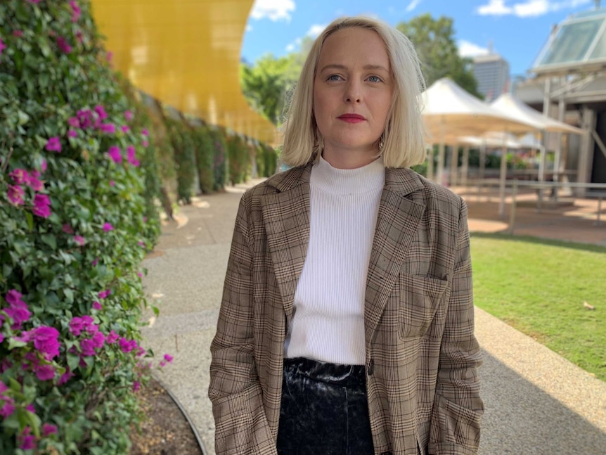 Claudia Levi stands next to a flowering hedge in a park on a sunny day.