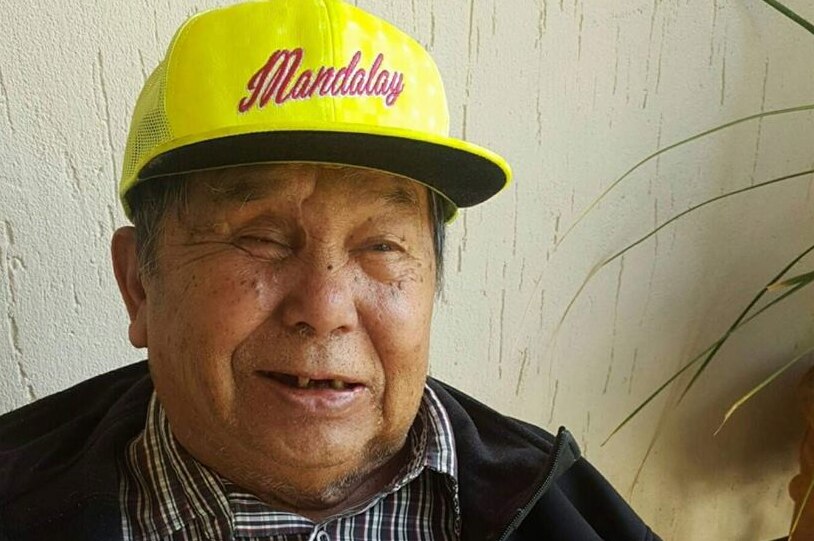 An elderly man - originally from Myanmar - smiles while wearing a yellow hat.