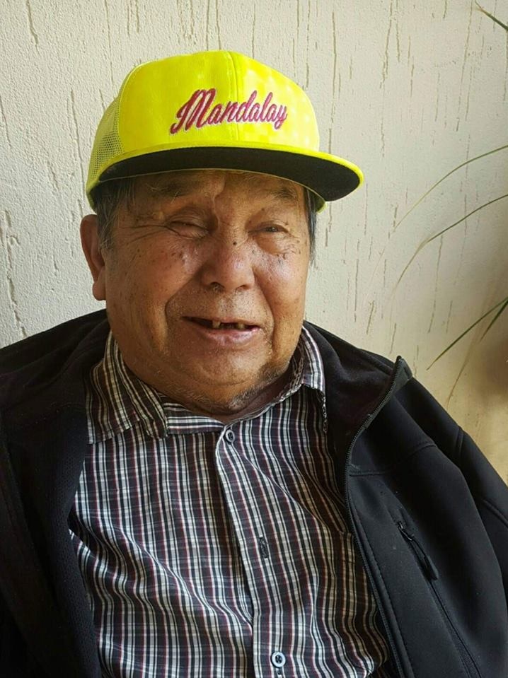 An elderly man - originally from Myanmar - smiles while wearing a yellow hat.