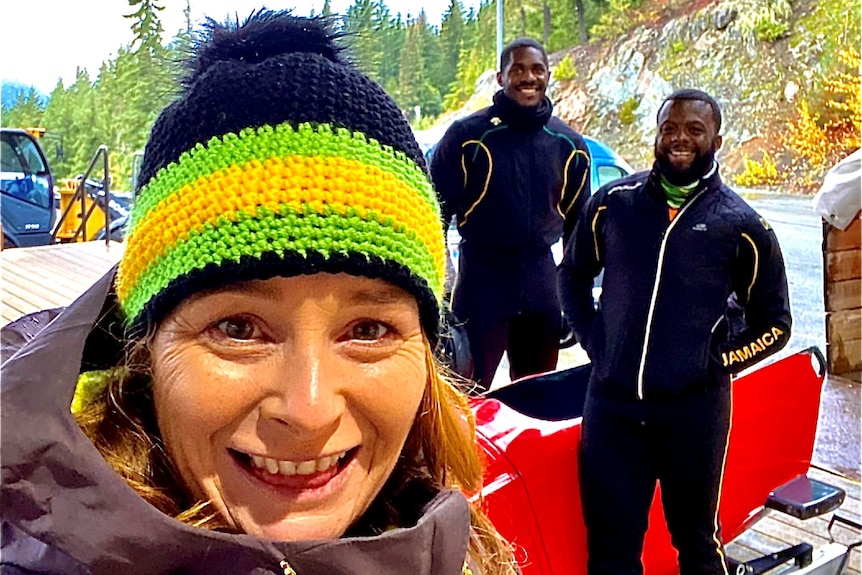 Lady close to the camera witha beanie, smiling with two athletes behind her standing at a bobsled