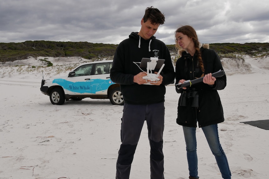 They stand on a beach with their 4WD behind them, holding a drone