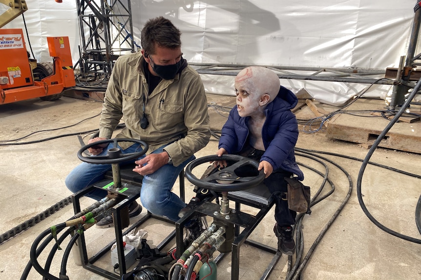 Man and teenage with dwarfism sit on a skeleton of a car on a movie set