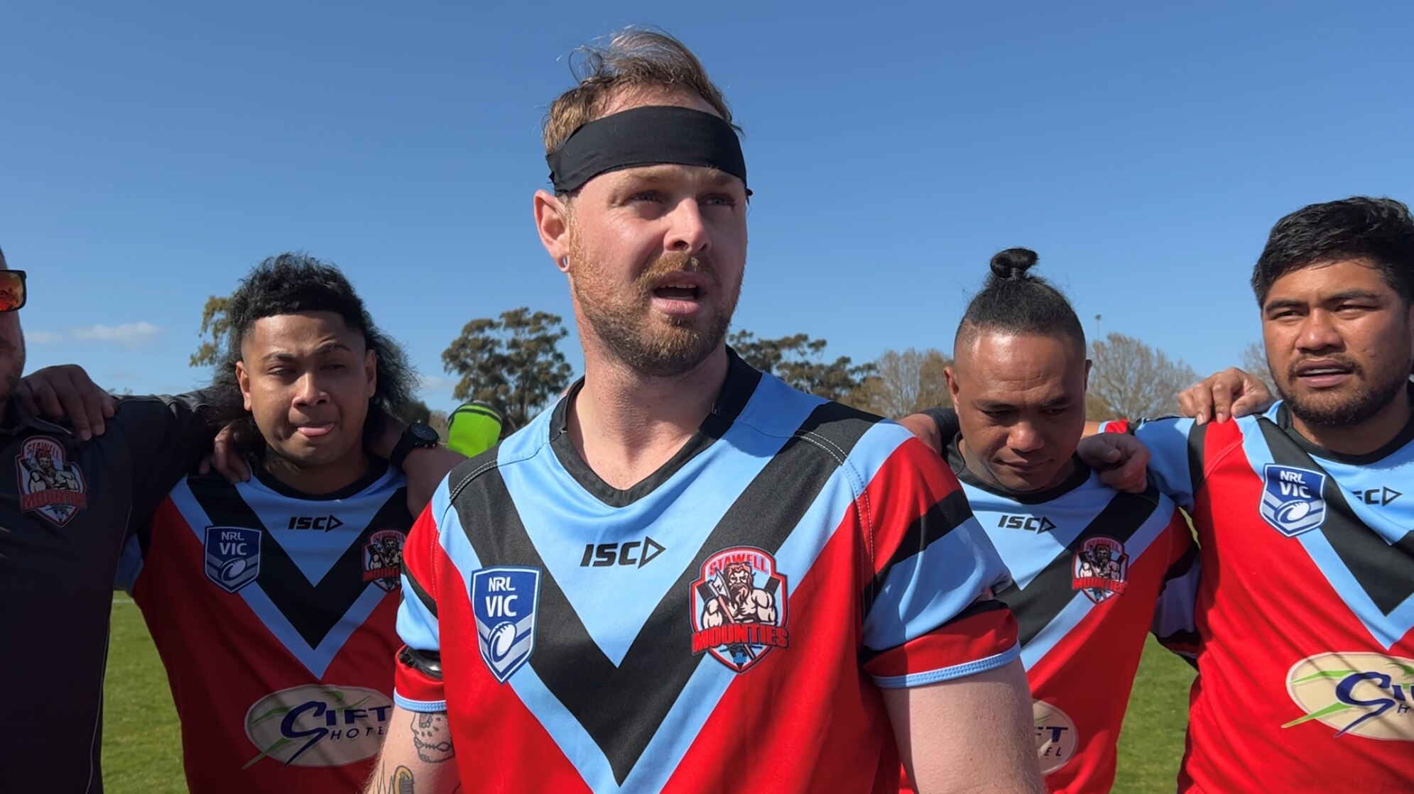 man with black headband and blue and red jersey addresses teammates in a huddle.
