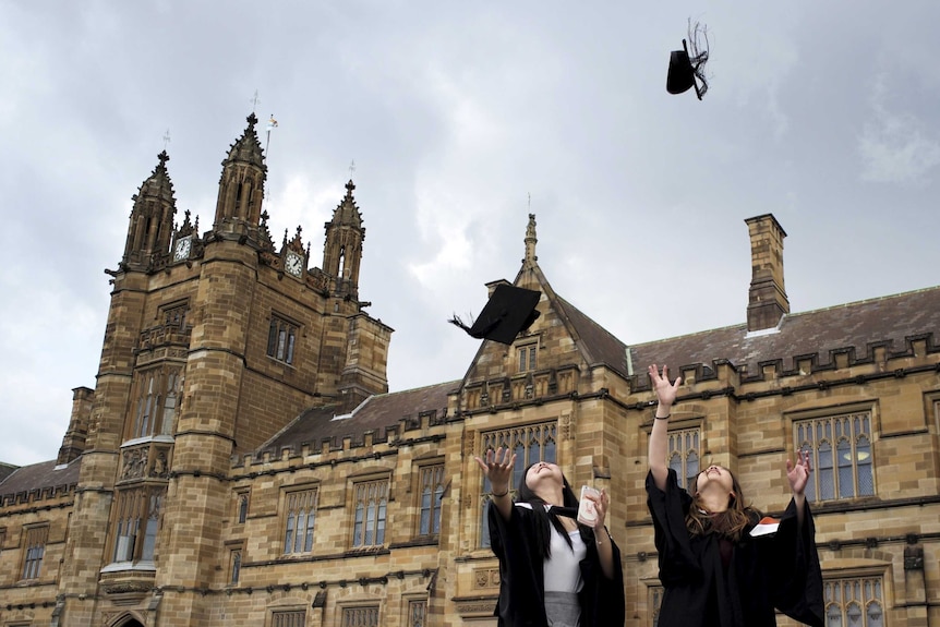 Two students in graduation gowns jump and throw their caps into the air outside a historic-looking building.