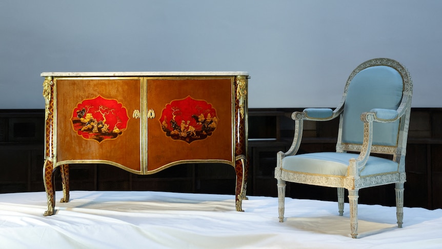 A chest of drawers with a gold border and painted design sits next to a grey chair. 