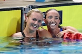 Two Australian female swimmers are together in a pool on either side of a lane rope smiling at the camera after a big race.