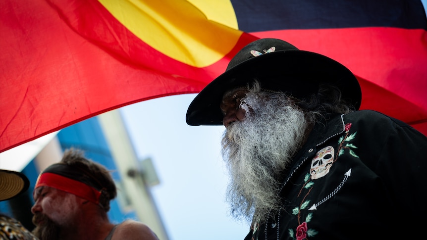 An Indigenous man standing in front of an Aboriginal flag.