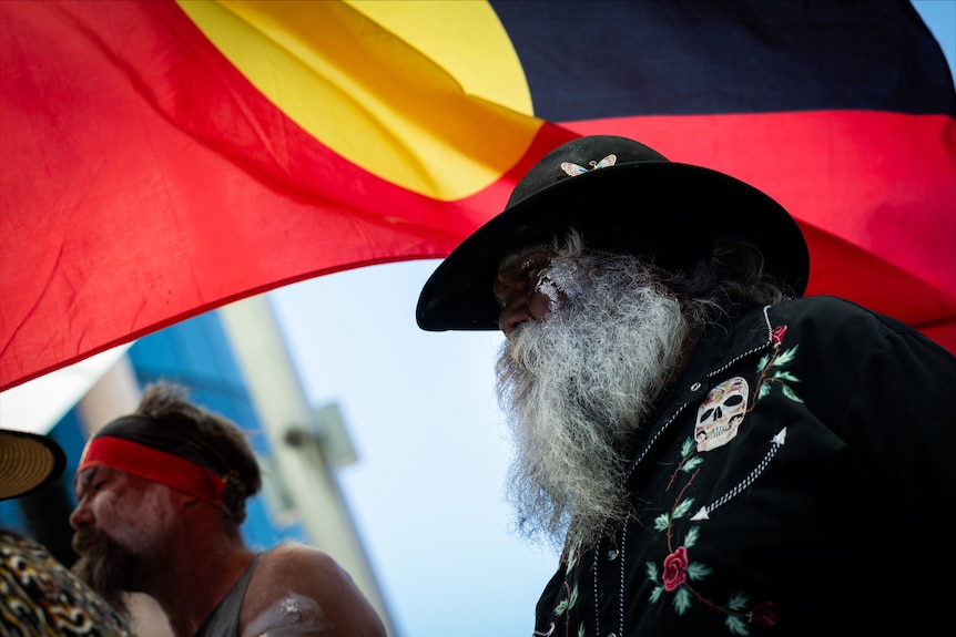 An Indigenous man standing in front of an Aboriginal flag.