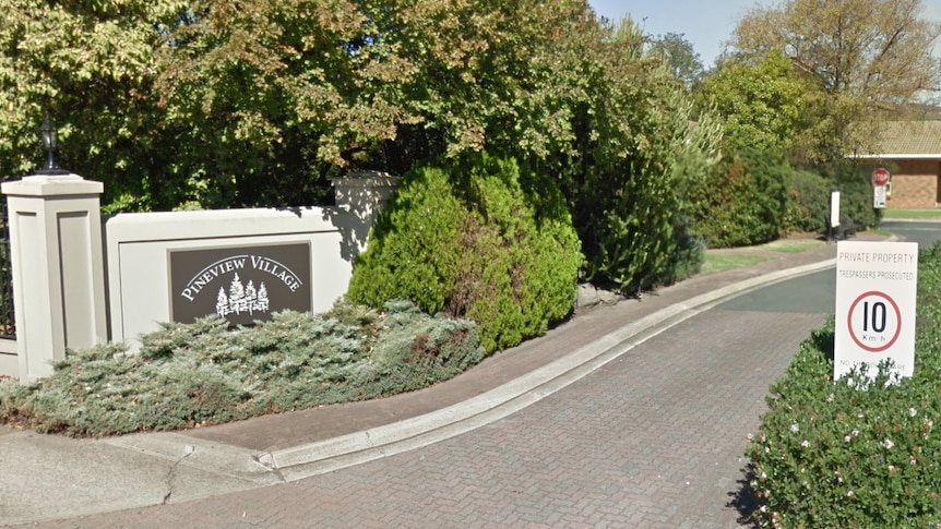 A sign saying Pineview Village placed next to a road which leads to a brown brick house