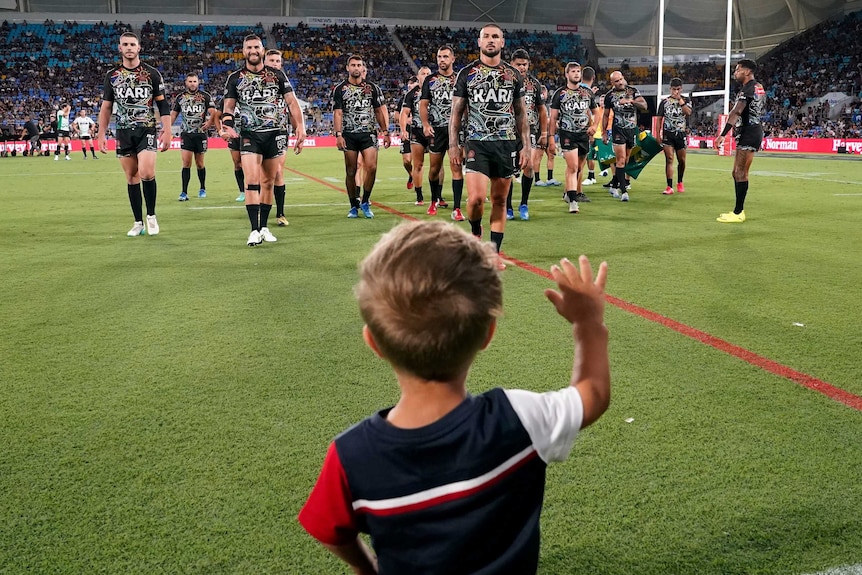 Quaden Bayles, seen from behind, waves to the Indigenous All Stars team as they warm up.