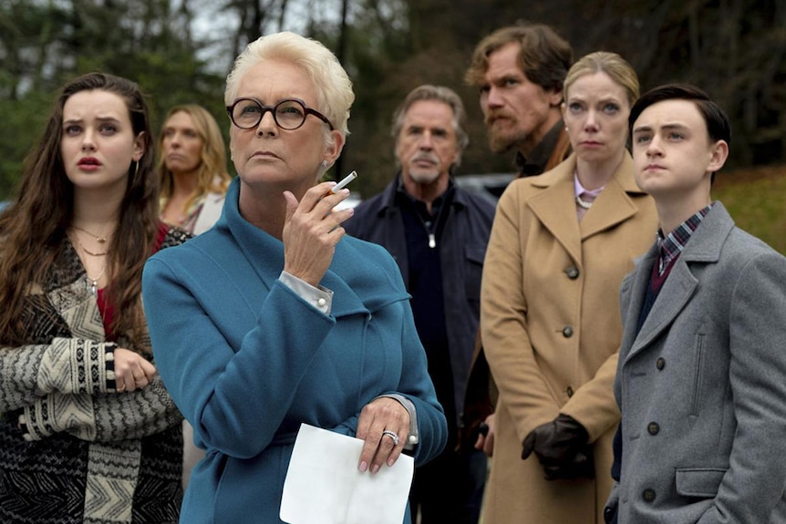 Jamie Lee Curtis in spectacles and teal wool coat holds cigarette and stands with a six others outdoors on a cold overcast day.